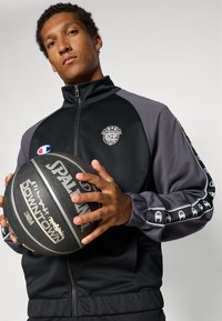 Jaunas vyras dėvi juodą ir pilką užtrauktuku užsegamą striukę, laiko juodą Spalding krepšinio kamuolį su baltais žymėjimais.