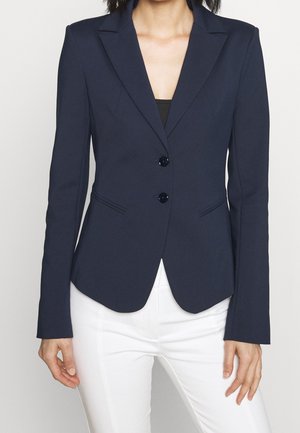 Kvinde iført en tætsiddende mørk marineblå blazer med to knapper over en sort top og hvide bukser, stående med armene afslappet ned langs siden.