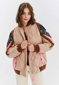 Přefouknutá bombera v světle béžové barvě s vzorovanými rukávy, růžovými akcenty a žebrovanými manžetami. Disponuje zipovým uzávěrem a dekorativními třásněmi.