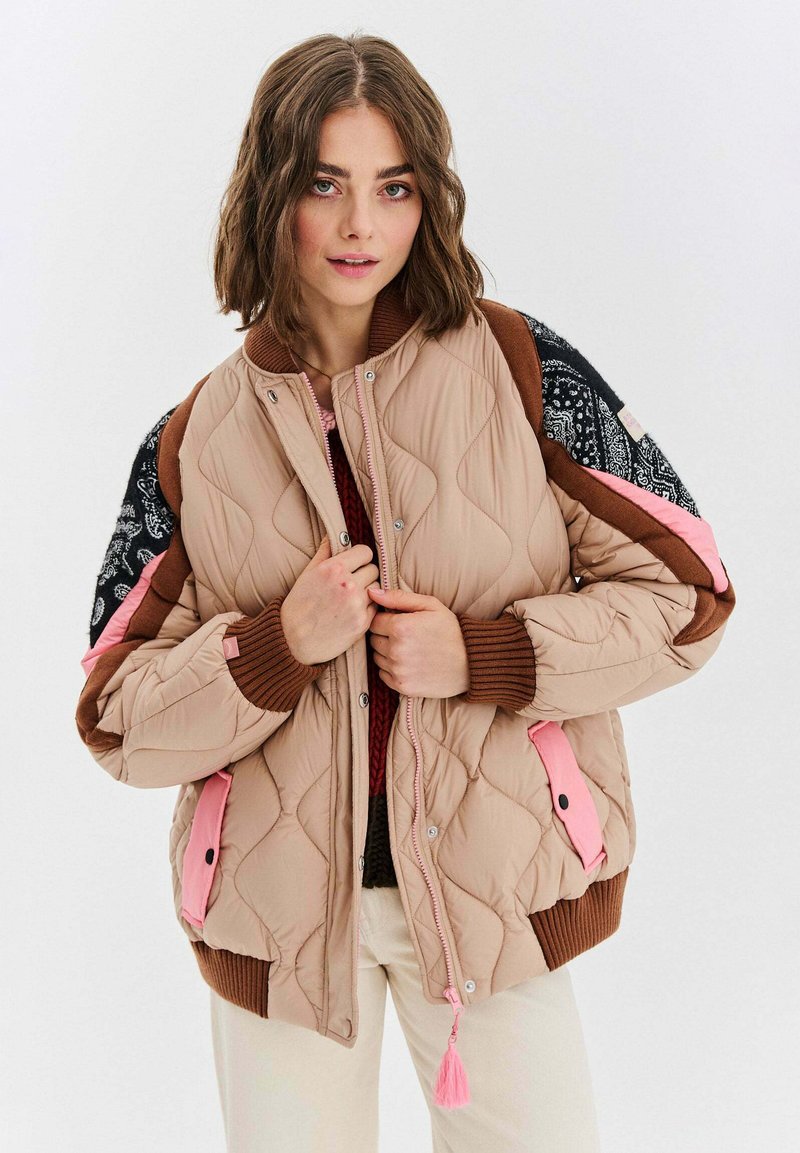Přefouknutá bombera v světle béžové barvě s vzorovanými rukávy, růžovými akcenty a žebrovanými manžetami. Disponuje zipovým uzávěrem a dekorativními třásněmi.