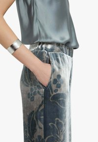 Top in raso grigio abbinato a pantaloni in velluto con motivo floreale nei toni del blu e del grigio, tasche laterali e braccialetto a polsino argento al polso.