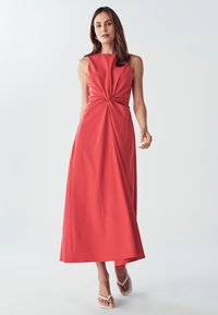 Korálové bezramenné šaty s uzlovým detailom v páse, splývavá sukňa a hladká textúra. V kombinácii s bielymi sandálmi.