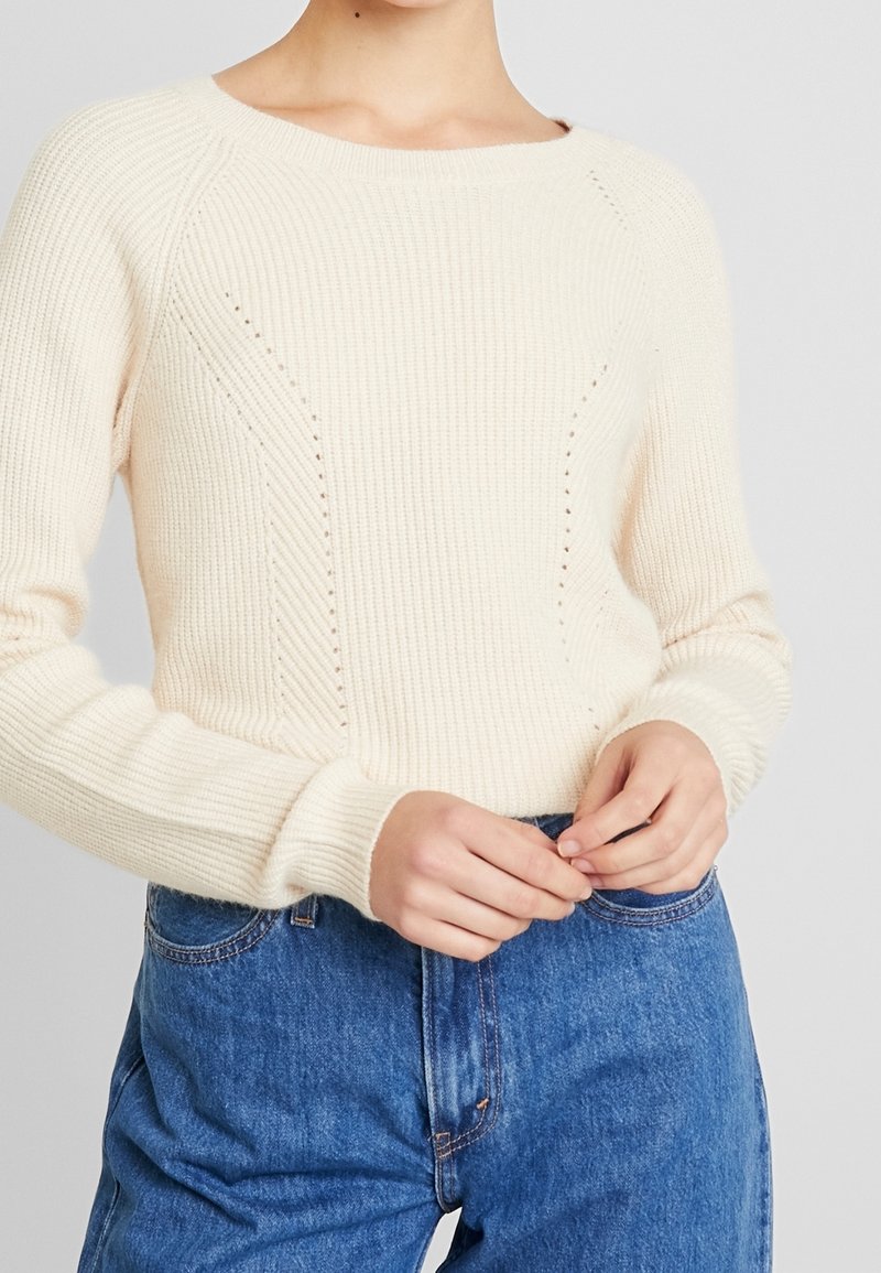 Cream-colored ribbed sweater with a wide neckline, featuring diamond-shaped stitch details and fitted cuffs, worn with high-waisted blue jeans.
