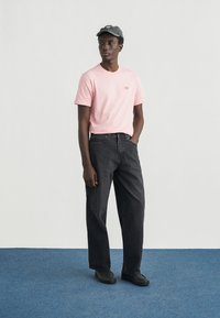 Ροζ κοντομάνικο μπλουζάκι με μικρό λογότυπο, μαύρο τζιν, γκρι καπέλο και μαύρα παπούτσια. Μεμονωμένη φιγούρα σε μπλε χαλί μπροστά σε λευκό φόντο.