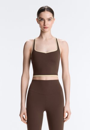 Mladá žena s uhladenými vlasmi, nosiaca hnedý krátky top a zodpovedajúce legíny s vysokým pásom, na jednoduchom svetlom pozadí.