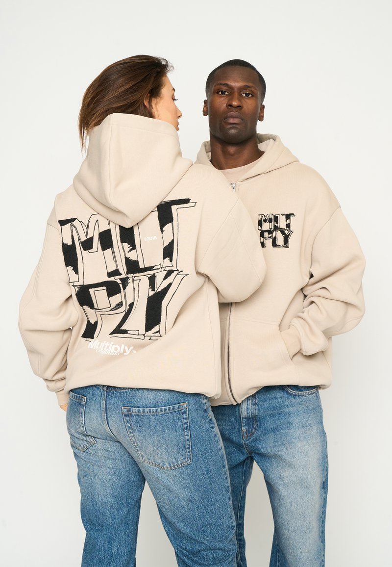 Las sudaderas beige con cremallera cuentan con estampados gráficos oversize en la parte trasera y delantera. La tela parece suave, con un ajuste relajado y bolsillos tipo canguro.