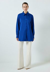 Cămașă albastră cu mânecă lungă și nasturi, având guler și o textură fină. Asortată cu pantaloni largi de culoare crem și tocuri bej.