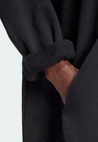 Svart stickad plagg med rullad ärm, som visar en mjuk textur och en löst sittande passform. En hand syns, vila nära en ficka.