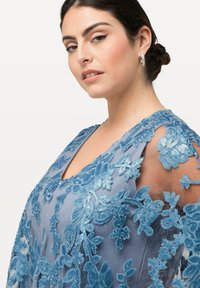 Spetsklänning i ljusblått med intrikata blommönster och paljetter. Genomskinliga ärmar och en V-ringning framhäver dess elegans och textur.