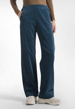 Persoon draagt blauwe brede ribfluwelen broek, beige sneakers en een bruin top met lange mouwen, staat met handen in de zakken.