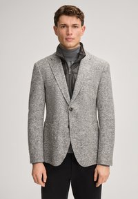 Grauer gewebter Blazer mit strukturiertem Muster, ausgestattet mit zwei Fronttaschen und einem Ein-Knopf-Verschluss, übereinander getragen mit einer schwarzen Jacke mit Reißverschluss.