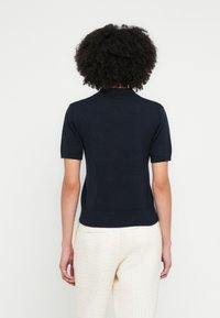 Sötétkék rövid ujjú kötött felsőrész bordázott gallérral és szegéllyel, sima textúrával és testhezálló fazonnal, világos színű, texturált nadrággal párosítva.