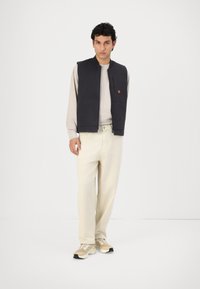Navy sleeveless vest with front zip and pockets, worn over a light beige long-sleeve shirt, paired with cream trousers and beige sneakers.