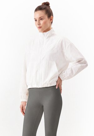 Donna con chignon che indossa una giacca bianca con zip e leggings grigi, in piedi con una mano sul fianco su sfondo bianco.