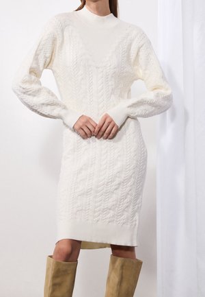 Woman wearing a white cable knit sweater dress with long sleeves and beige knee-high boots, standing with hands clasped in front.