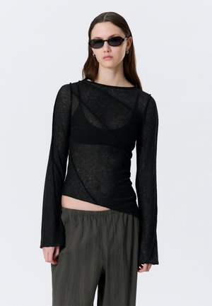 Donna che indossa una maglia nera trasparente a maniche lunghe con design asimmetrico, occhiali da sole scuri e pantaloni larghi verde oliva su sfondo grigio uniforme.