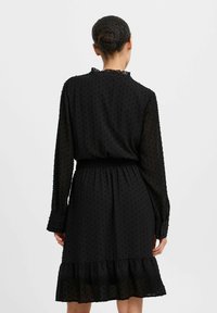 Robe noire en tissu transparent avec une texture à pois, manches longues et ourlet à volants. Présente une taille élastique et un col à volants.
