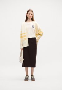 Gilet crème avec des rayures jaunes, t-shirt blanc avec logo, jupe midi marron, baskets grises et petit sac à bandoulière clair.