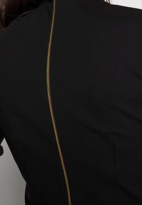 Vêtement noir avec une fermeture éclair dorée visible courant verticalement le long du dos. Texture de tissu lisse avec des lignes épurées et une coupe ajustée.