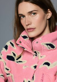 Roza flis jakna s kremnimi in mornarsko modrimi vzorci. Visok ovratnik, zapenjanje na pritrdilne gumbe in detajl žepa z zadrgo. Mehka, teksturirana tkanina.