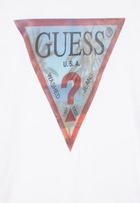 T-shirt blanc avec un graphique triangulaire coloré comportant le texte "GUESS U.S.A." et "WASHED JEANS," ainsi que les numéros 1201 et 1203.