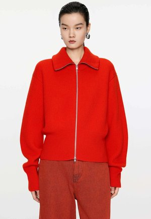 Red ribbed knit zip-up jacket with a wide collar and long sleeves. Features a silver zipper and a fitted waist, worn over matching red pants.