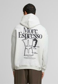 Lys grå hættetrøje med et sort grafik af en kaffemaskine med etiketter, herunder "More Espresso," "filter," og "vandtank."