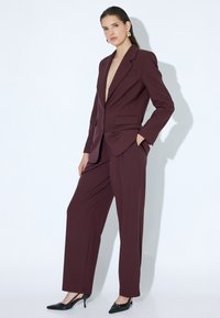 Costume ajusté bordeaux avec un blazer en V profond et un pantalon à larges jambes. Porté avec des talons pointus noirs, avec un tissu lisse et une coupe structurée.