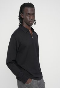 Μαύρο πόλο μπλουζάκι με μακριά μανίκια και φερμουάρ στον γιακά, φτιαγμένο από μαλακό ύφασμα. Διαθέτει κανονικά μανσέτες και άνετη εφαρμογή. Διακριτικές λεπτομέρειες ραφής.