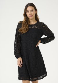 Schwarzes Spitzenkleid mit komplizierten Mustern, langen Ärmeln und einem gewellten Ausschnitt. Die Länge reicht bis über das Knie und hat eine lockere Passform.