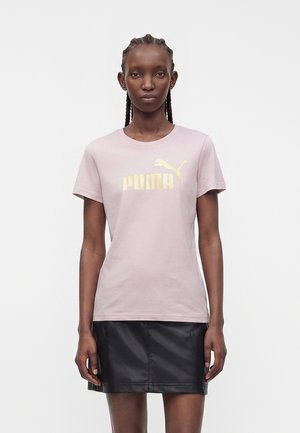 Kvinde med flettet hår iført en lyserød Puma T-shirt med gyldent logo og et sort lædernederdel, stående mod en hvid baggrund.