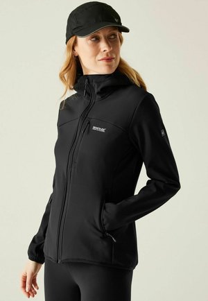 Woman wearing black softshell jacket with zipped pockets and black cap, looking to her left against plain background.