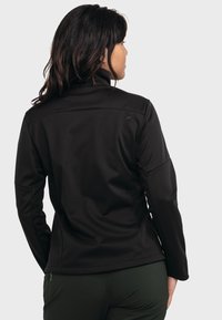 Schwarze Softshelljacke mit hohem Kragen, tailliertem Schnitt und langen Ärmeln. Besitzt dezentes Branding im oberen Rückenbereich. Glatte Textur.