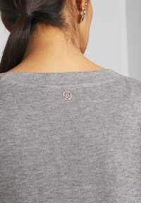 Grå stickad tröja med rund halsringning, med en liten, metallisk logotypaccent på baksidan. Slät textur med en enkel design.