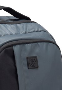 Grå rygsæk med en quiltet tekstur, sorte detaljer og et logo-patch. Har polstrede remme og en organiseret ruminddeling.