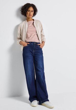 Hellbeige Bomberjacke, pink gestreiftes T-Shirt, weite dunkelblaue Jeans, weiße Sneaker mit strukturierten Akzenten, stehend vor einem weißen Hintergrund.