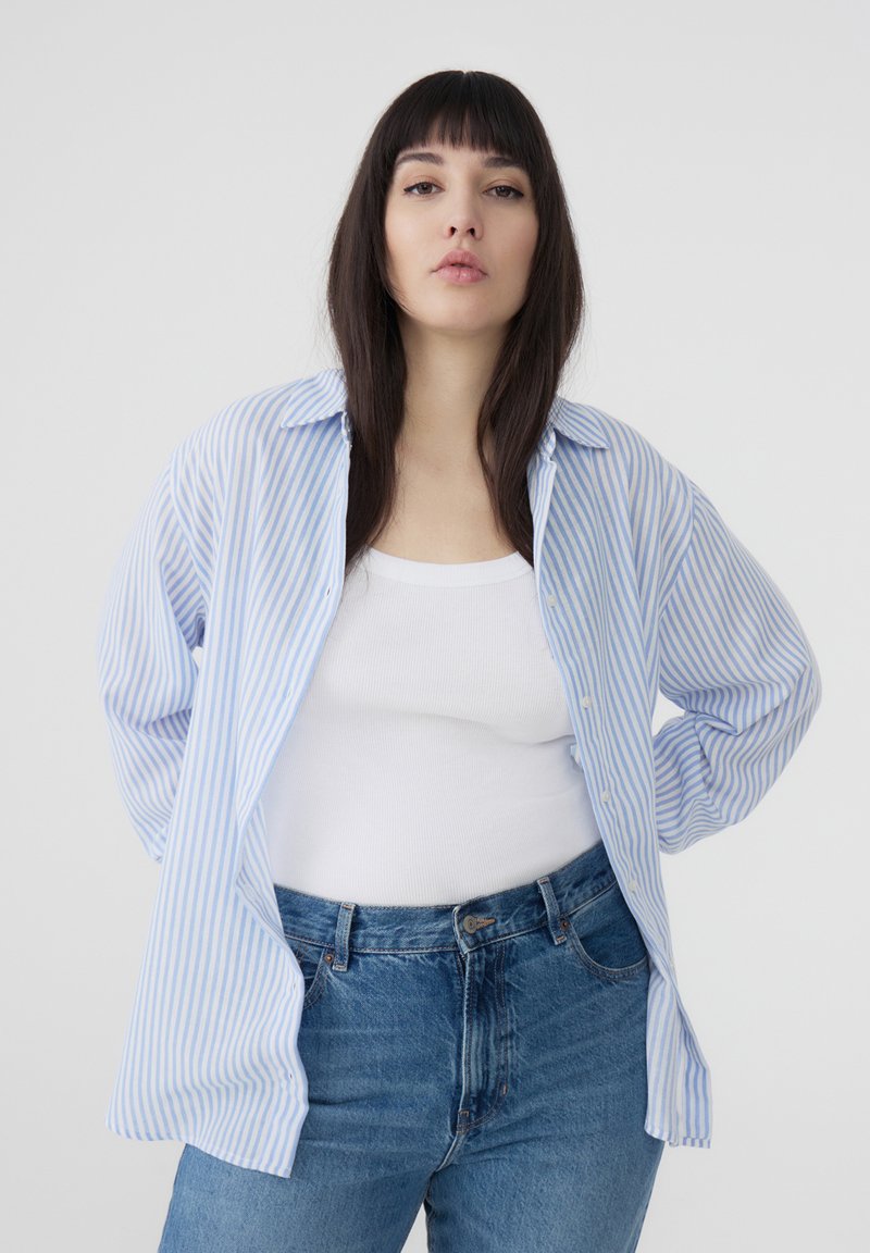 Camicia button-up a righe blu chiaro e bianco, con maniche lunghe, indossata sbottonata sopra un top bianco. Abbinata a jeans di denim blu.