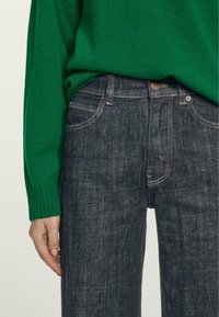 Jeans di denim blu scuro con taglio dritto, caratterizzati da cuciture a contrasto, tasche frontali e chiusura con bottone. Abbinati a un maglione verde.