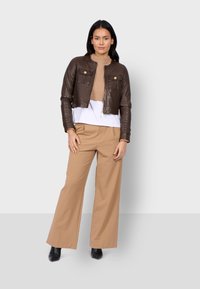 Giacca cropped in pelle marrone con due tasche frontali, abbinata a un dolcevita beige a coste e pantaloni beige larghi, completata da stivali neri alla caviglia.