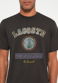 Černé bavlněné tričko s logem "LACOSTE" v zelené a oranžové barvě, se grafikou krokodýla a textovými detaily níže. Klasický střih s kulatým výstřihem.