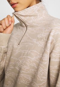 Beigen värinen poolopaita, jossa on puolivetoketjullinen kaulus, hienovarainen sävyeroilla toteutettu abstrakti kuvio ja pieni kirjailtu "Levi's"-logo rinnassa.