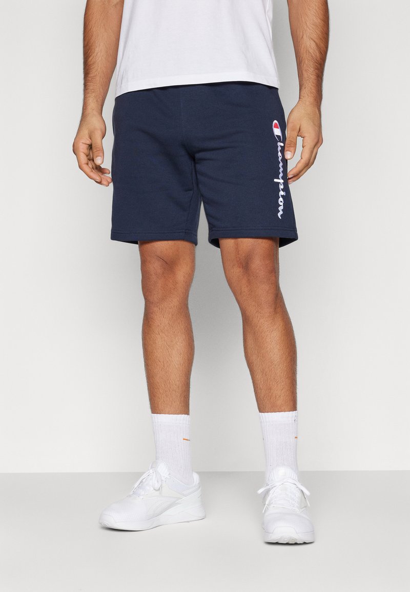 Shorts de algodón color marino con un acabado texturizado y el logo blanco de "Champion" en el lateral, combinados con zapatillas deportivas blancas y calcetines de longitud media.