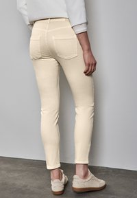 Pantalones vaqueros skinny de color beige claro con una textura suave, dos bolsillos traseros y una cintura de altura media, combinados con zapatillas de deporte beige texturizadas.