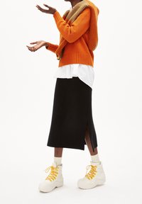 Personne portant un pull orange avec une écharpe assortie, une chemise blanche, une jupe midi noire fendue sur le côté, et des bottes blanches montantes à lacets jaunes.