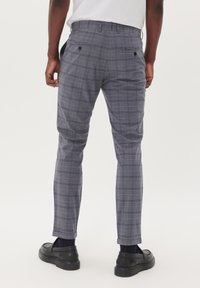 Pantalones de cuadros grises con un corte ajustado, fabricados en tejido ligero. Presentan bolsillos traseros con cierres de botón y puños enrollados. Calzado negro visible.