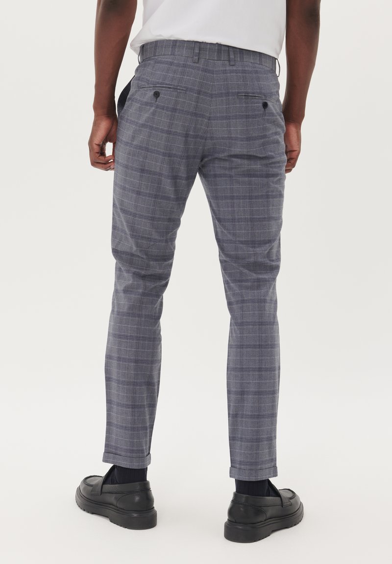 Grijze geruite broek met een slim fit, gemaakt van lichte stof. Voorzien van achterzakken met knoopsluitsels en opgerolde manchetten. Zwarte schoenen zichtbaar.