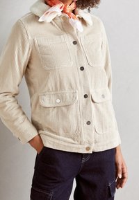 Chaqueta de pana en color beige claro con cuello de felpa, que cuenta con dos bolsillos frontales, botones de presión y textura acanalada.
