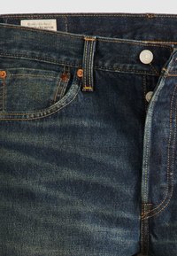 Jeans de mezclilla de color azul oscuro con desgastes visibles, remaches de cobre, costuras amarillas-naranjas y una etiqueta de tela marrón en la cinturilla.