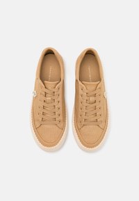 Tan canvas sneakers with round toe, textured fabric, beige laces, and a contrasting white rubber sole. Features a small metallic logo detail.