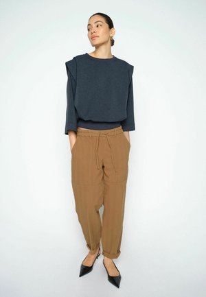 Vrouw staand met handen in de zakken, draagt een donkerblauw shirt met lange mouwen, losse bruine broek en zwarte puntige schoenen tegen een effen witte achtergrond.